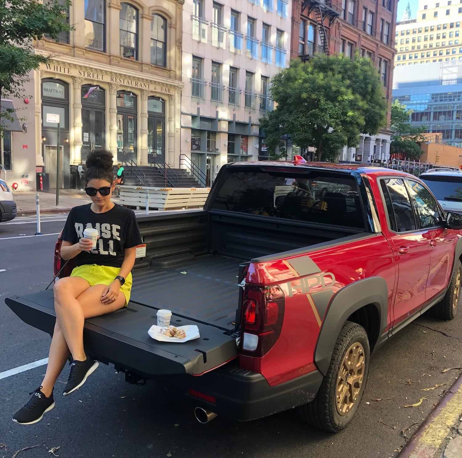 Woman sits on the tailgate of a used 2021 Honda Ridgeline compact pickup truck, buildings visible in the background.