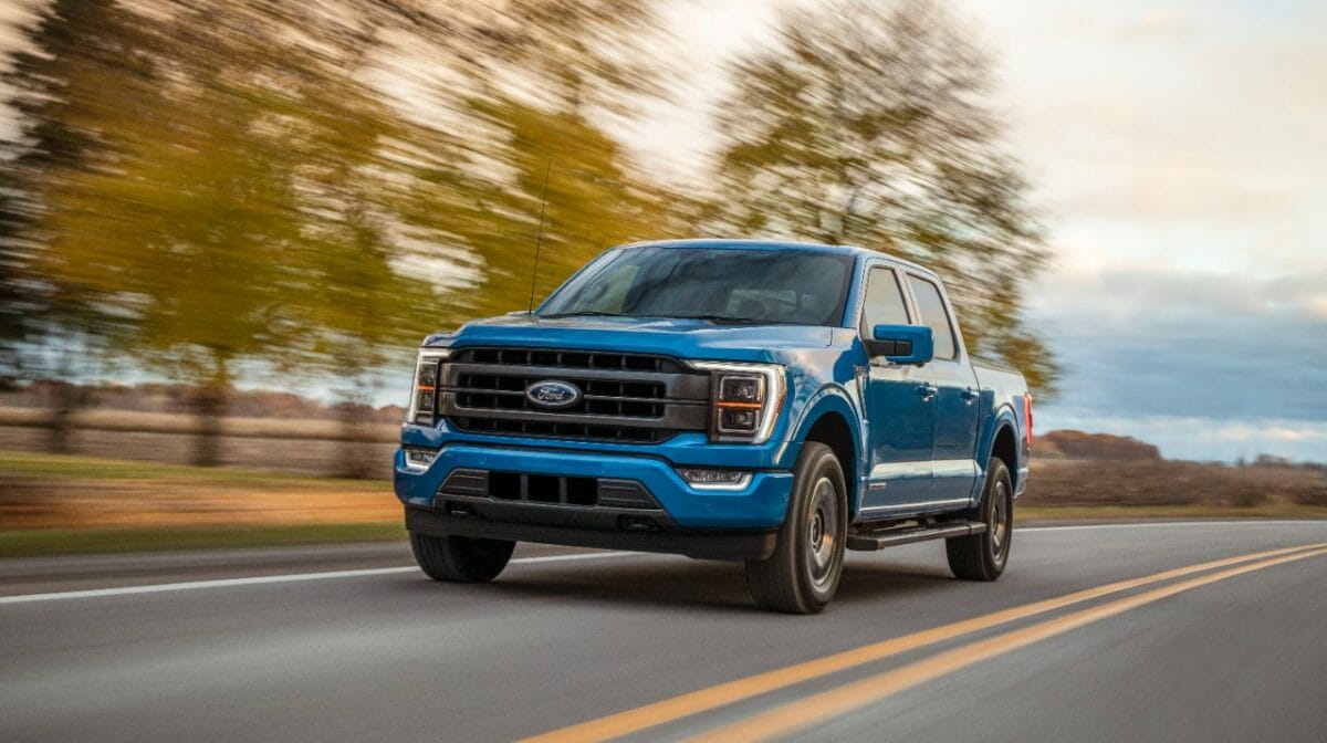 A blue Ford PowerBoost hybrid F-150 pickup truck driving along with tree's visible in the background.