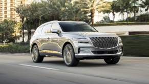 A silver 2024 Genesis GV80 drives past palm trees and high-rise condo buildings
