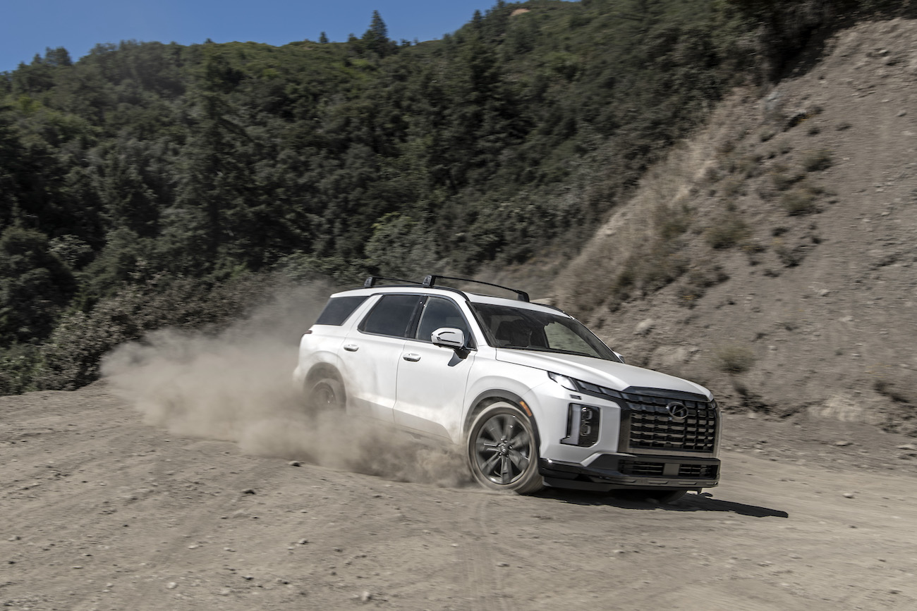 A white 2024 Hyundai Palisade driving on a dusty road. The Hyundai Palisade price is a perk compared to the Infiniti QX60