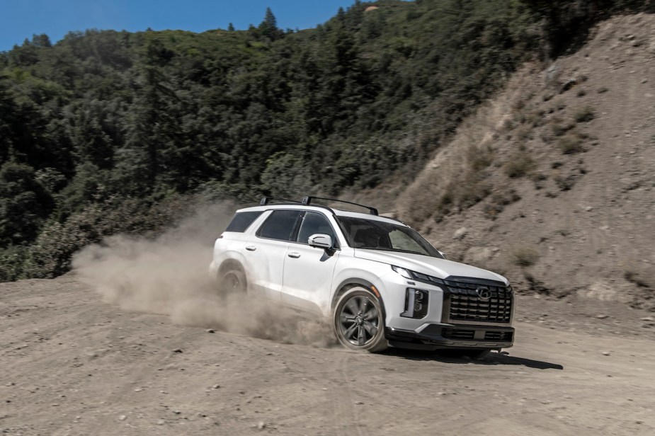 A white 2024 Hyundai Palisade driving on a dusty road. The Hyundai Palisade price is a perk compared to the Infiniti QX60