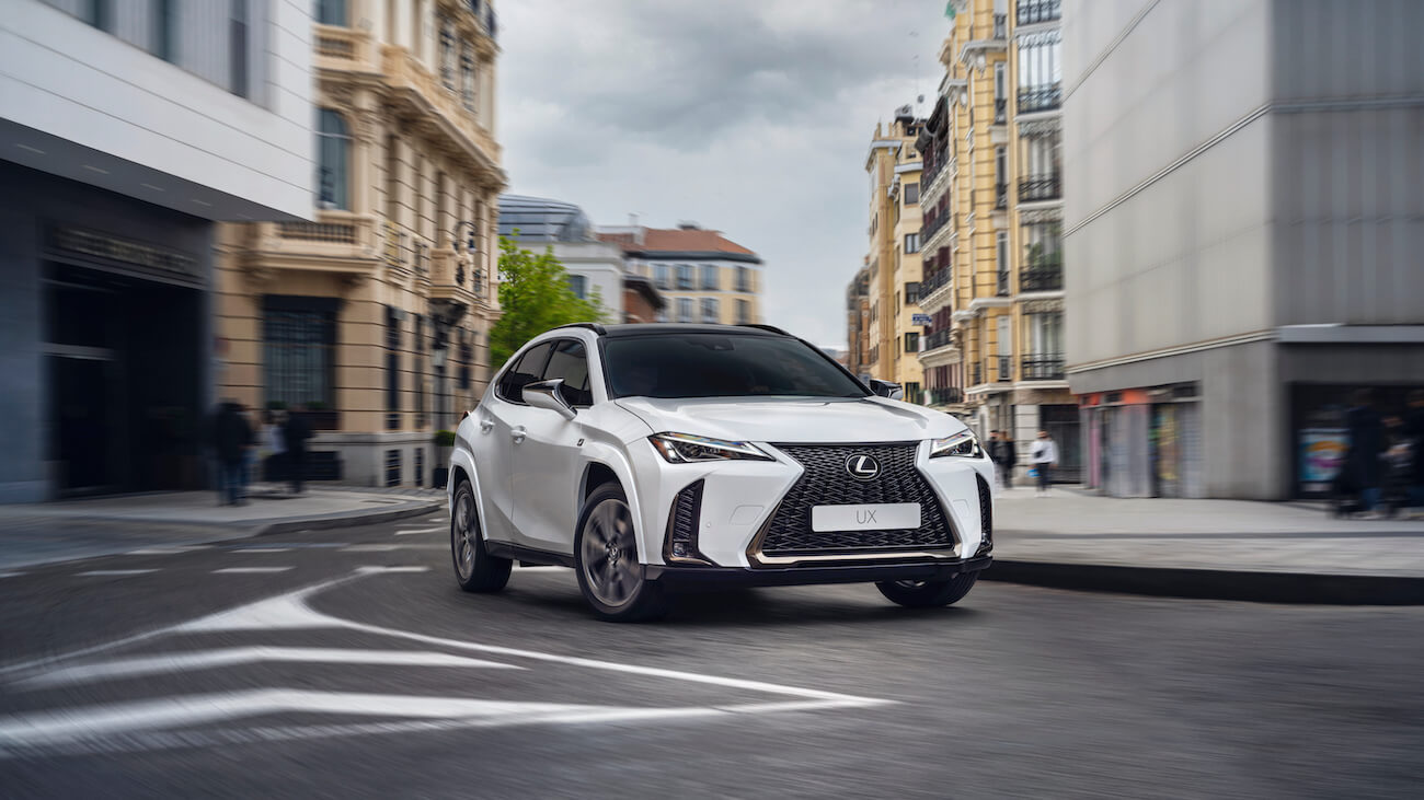 A white 2024 Lexus UX Hybrid taking a turn on a city street. The Lexus UX Hybrid price is one of its best selling points.