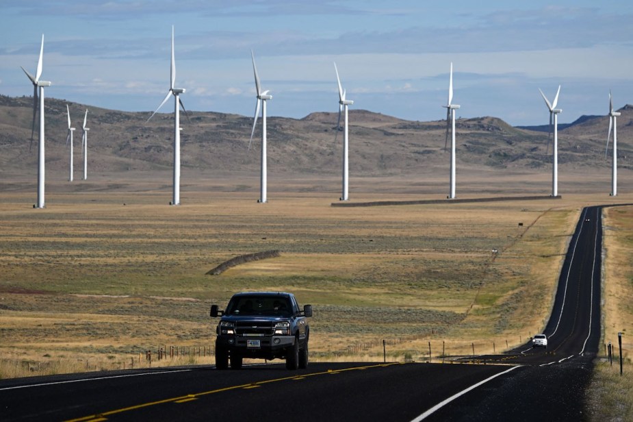 A Chevy Silverado pickup truck drives down a Wyoming road, wind turbines visible in the background.