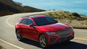 A red 2023 Genesis GV70 small luxury SUV is driving on the road.