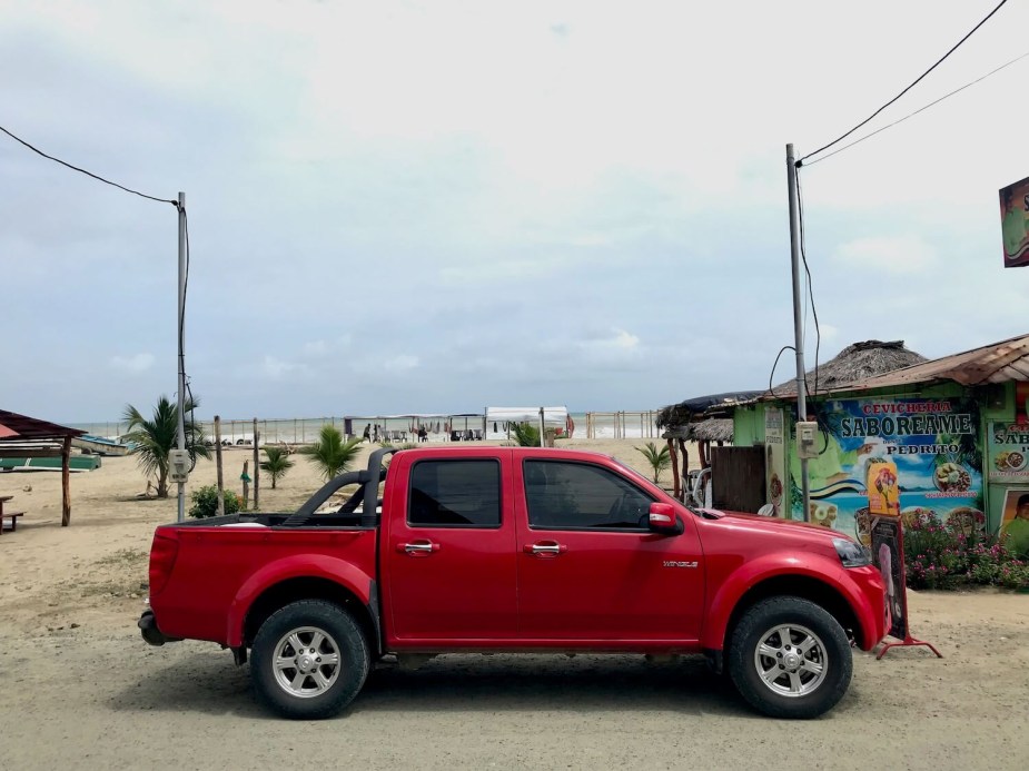 The side of a red, compact "Wingle" pickup truck parked on a beach in Ecuador, the ocean visible in the background.