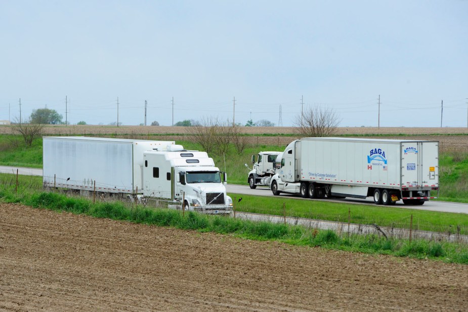 Three semi-truck tractor trailers drive in opposite directions along Indiana's I-74 interstate highway, unplanted farm fields visible in the foreground.