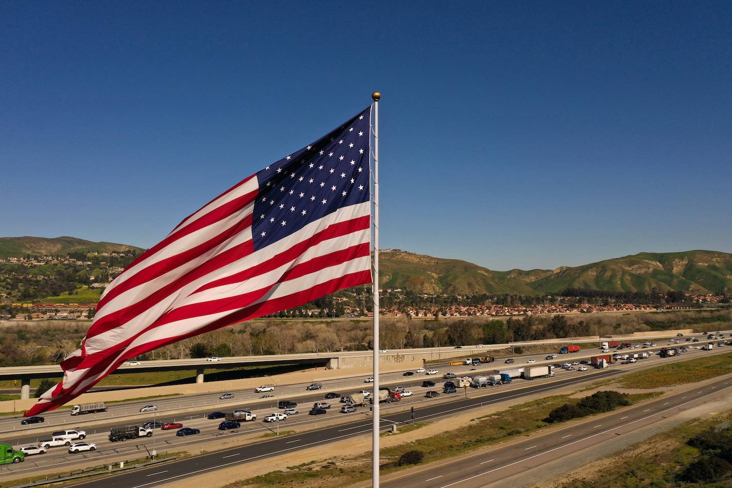 A string of cars and pickup trucks drive along a distant highway, the flag of the U.S.A. blowing in the foreground.