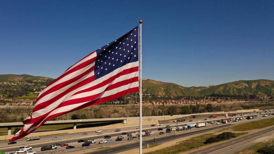 A string of cars and pickup trucks drive along a distant highway, the flag of the U.S.A. blowing in the foreground.