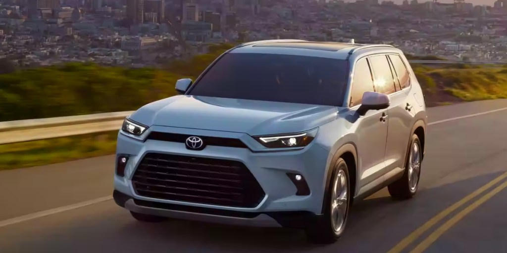 A white 2024 Toyota Grand Highlander midsize three-row SUV is driving on the road.