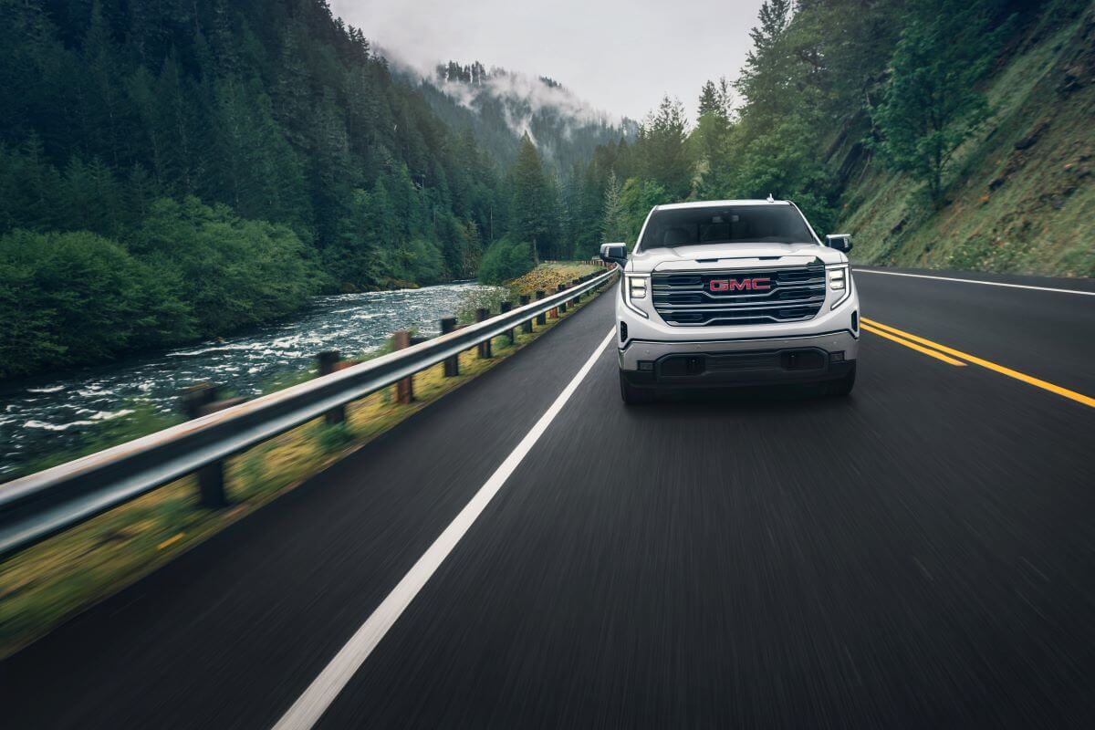 A 2024 GMC Sierra 1500 SLT Crew Cab Short Box 4WD full-size pickup truck model driving past a forest river