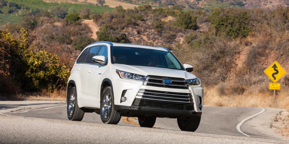 A white 2018 Toyota Highlander Hybrid midsize SUV is driving on the road.
