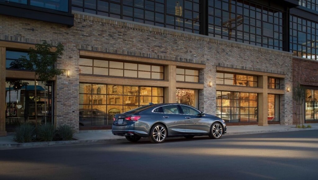 A gray 2020 Chevrolet Malibu sits next to a large building.
