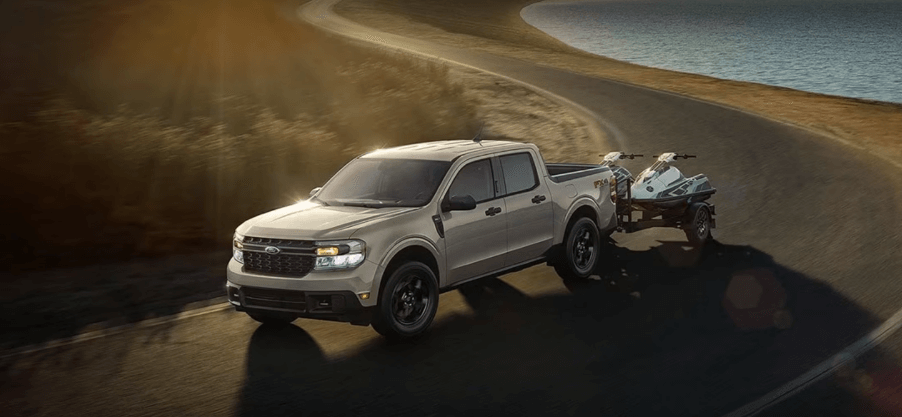 A 2024 Ford Maverick compact pickup truck model towing a pair of ATVs past a lake