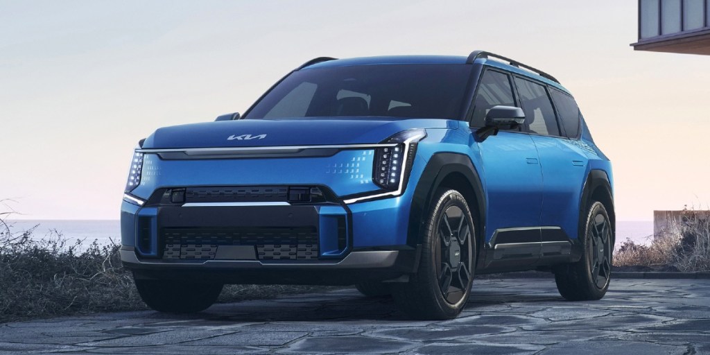 A blue 2024 Kia EV9 midsize electric SUV is driving on the road.