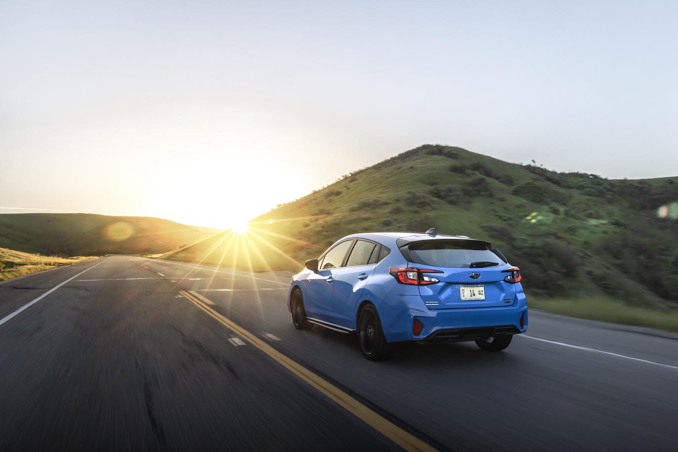 The 2024 Subaru Impreza RS trim level in blue driving into the sunrise on a road by a hill.