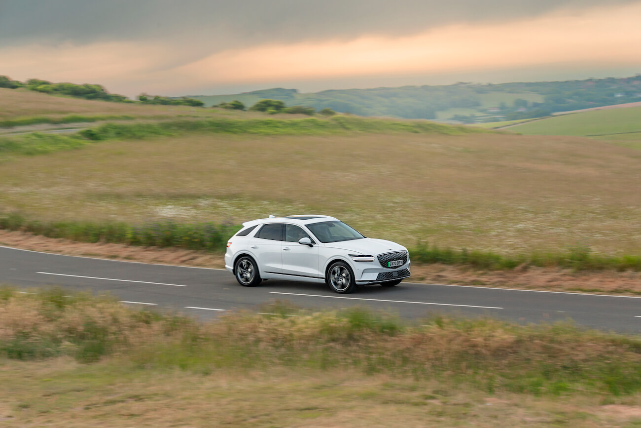A white Electrified Genesis GV70 driving along a winding road. Genesis service offers plenty of perks for owners.