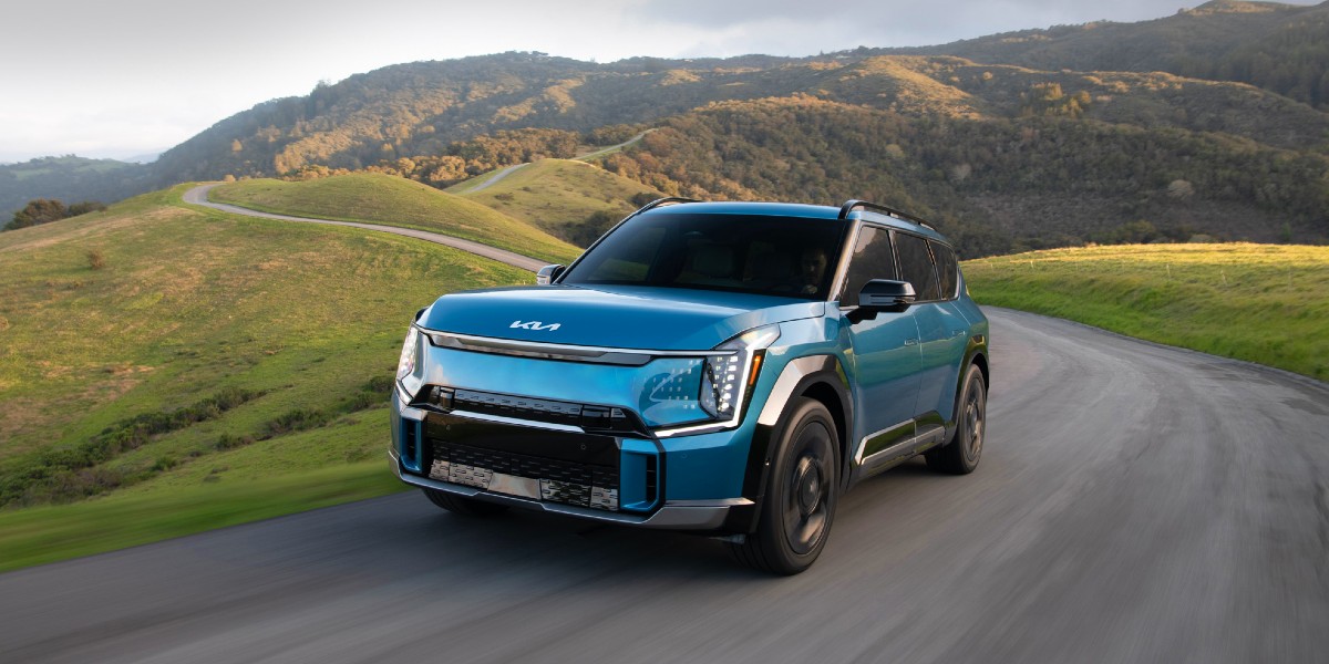 A blue 2024 Kia EV9 midsize electric SUV is driving on the road.