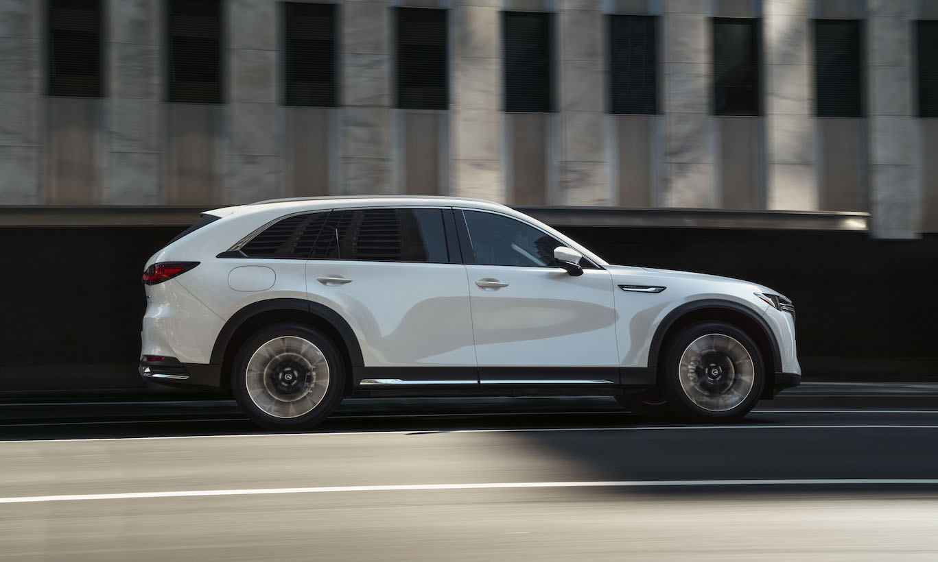 A white Mazda CX-90 driving down a city road. The CX-90 is the most expensive Mazda.