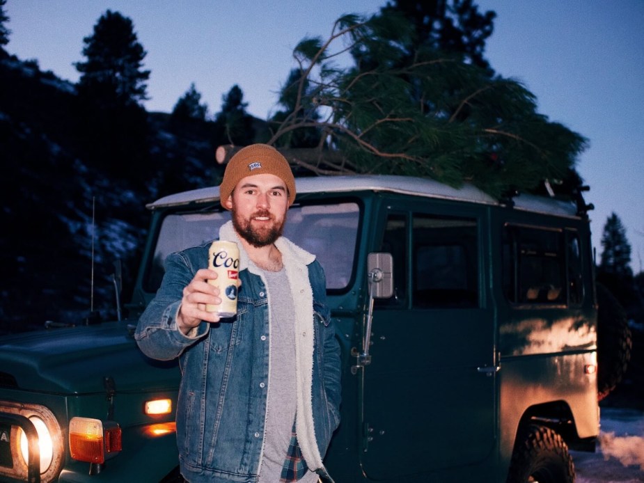 Man holds up a beer while standing in front of a Toyota FJ40 Land Cruiser SUV.
