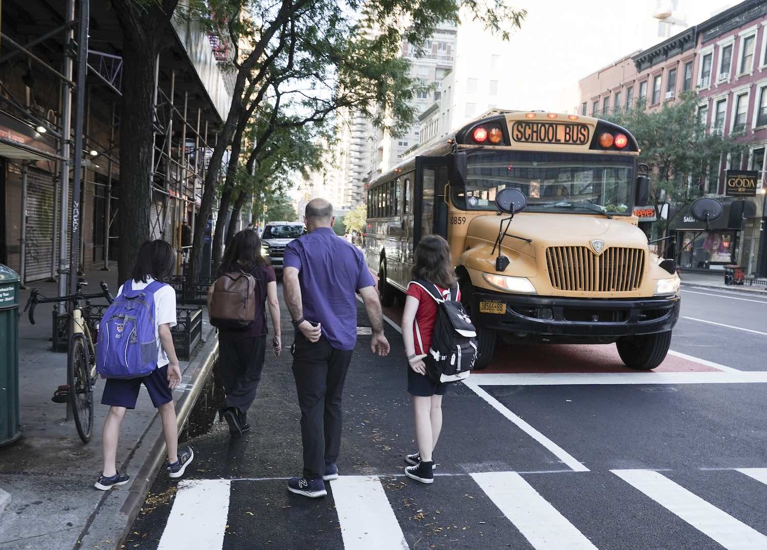 Students board a school bus on a city street in Manhattan New York, a grownup walking behind them.