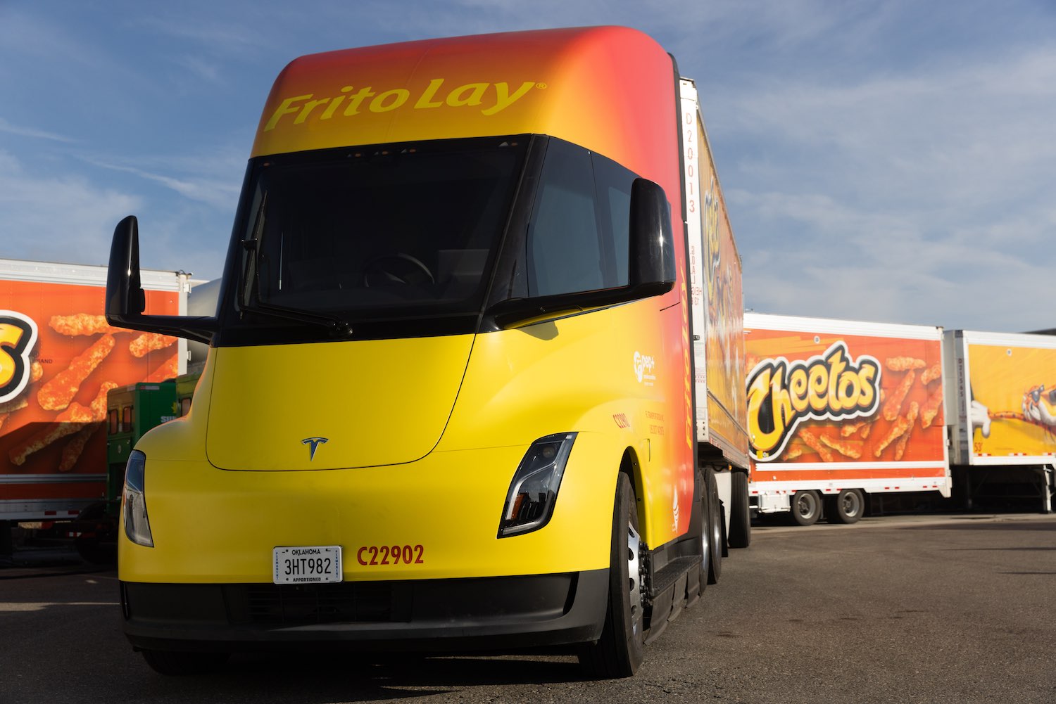 A bright yellow and orange Tesla semi truck tractor in front of a Cheetohs trailer in a parking lot.