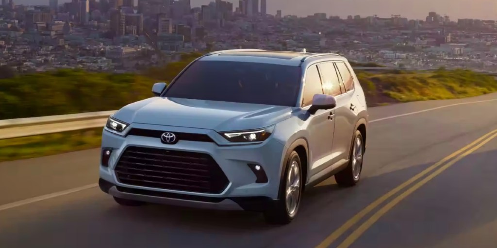 A white 2024 Toyota Gand Highlander Hybrid midsize three-row hybrid SUV is driving on the road. 