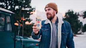 Man in a winter jacket holds up a beer, his Toyota SUV visible in the background.
