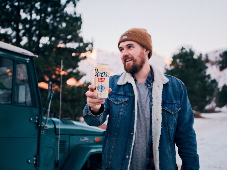 Man in a winter jacket holds up a beer, his Toyota SUV visible in the background.