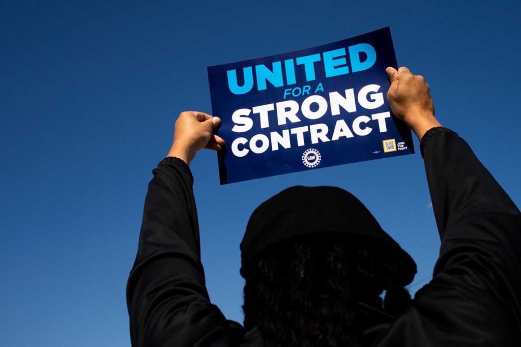 UAW worker holding up a picket sign while striking against Ford, GM, and Stellantis for better pay among record high profits
