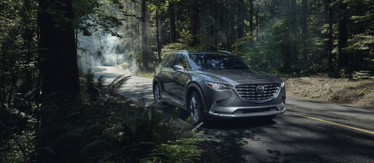 A 2023 Mazda CX-9 midsize SUV model parked on the side of a forest road shadowed by tree branches