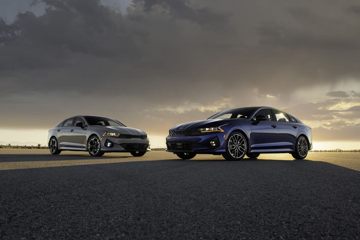 A pair of 2024 Kia K5 midsize sedan models parked on an asphalt lot and framed by a setting sun