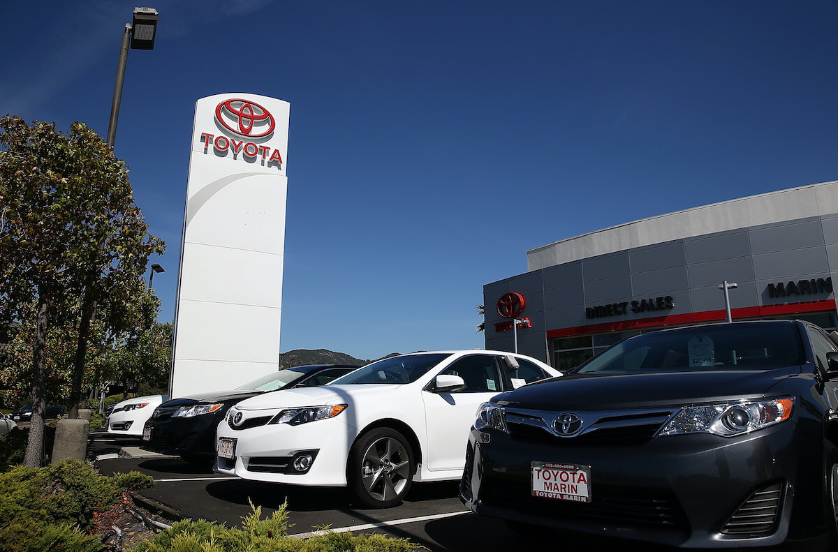 An expansive Toyota dealership lot under the clear blue sky, featuring rows of shiny new and pre-owned vehicles neatly arranged in orderly rows. The showroom building stands prominently at the center, with a sleek glass facade and the Toyota logo prominently displayed. Customers and sales staff can be seen inspecting cars, and colorful Toyota banners and flags flutter in the gentle breeze, adding a vibrant touch to the scene.