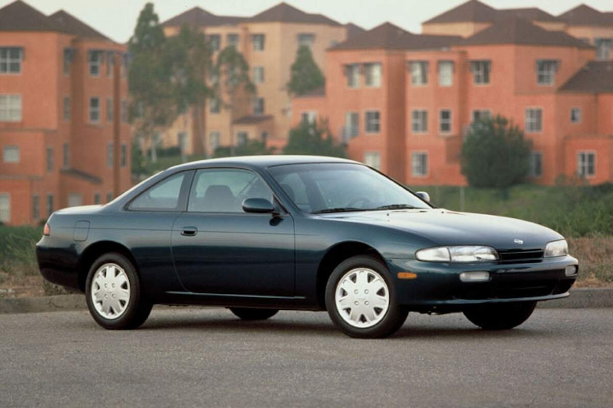 A dark-colored 1996 Nissan 240SX parked in front of orange buildings