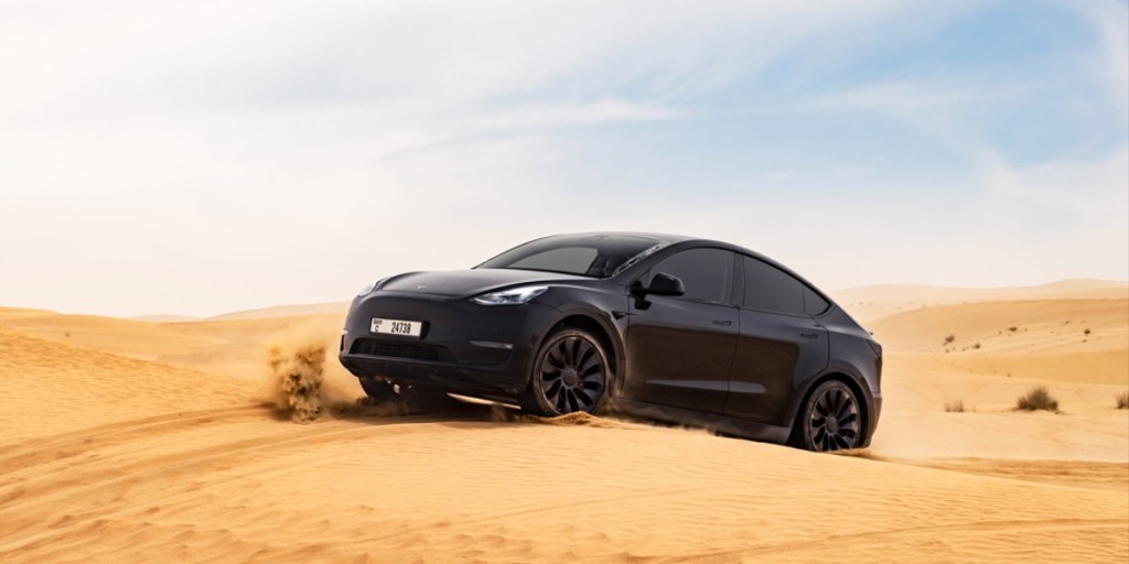 A black Tesla Model Y small electric SUV is driving in the sand.