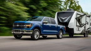 A blue 2024 Ford F-150 XLT pickup truck towing an RV up a road lined with trees.