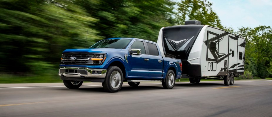 A blue 2024 Ford F-150 XLT pickup truck towing an RV up a road lined with trees.
