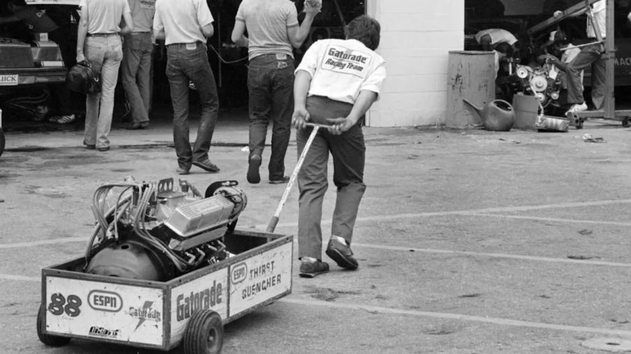 Black and white photo of a crew member pulling an old NASCAR V8 engine in a cart.