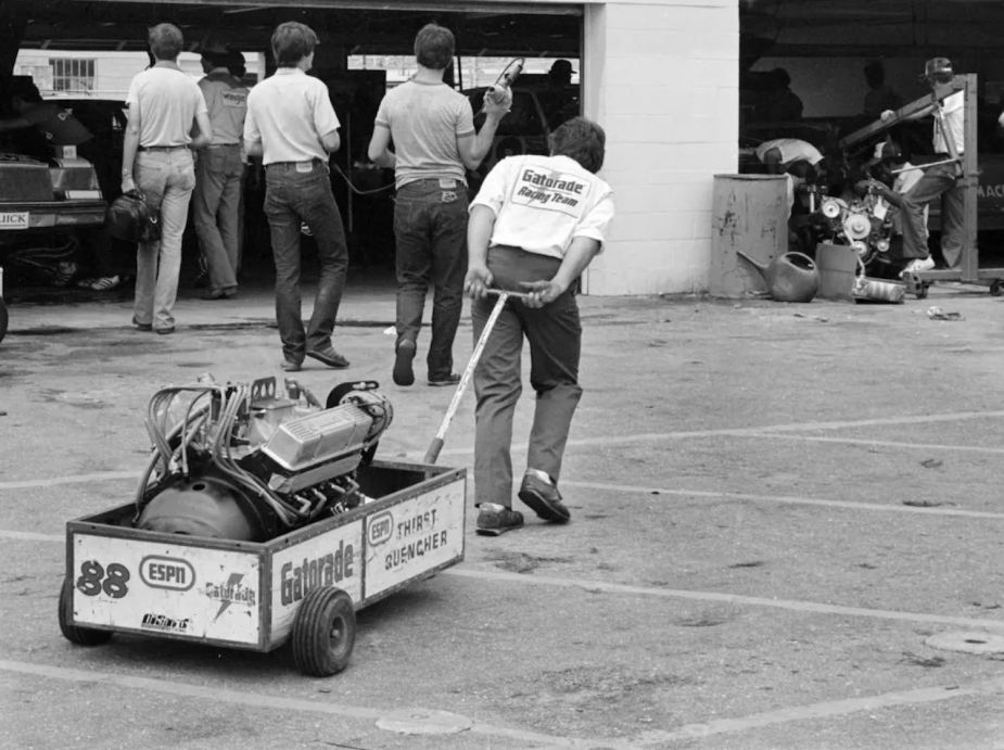 Black and white photo of a crew member pulling an old NASCAR V8 engine in a cart.