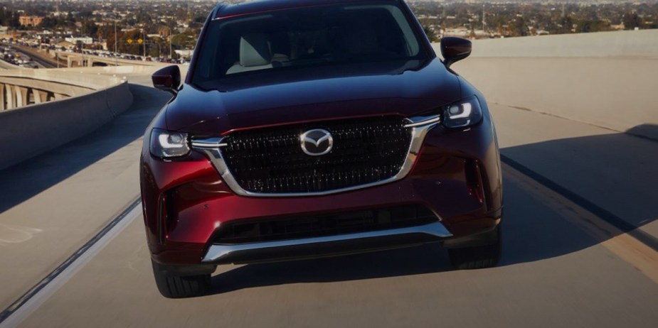 A red 2024 Mazda CX-90 midsize three-row SUV is driving on the road. 