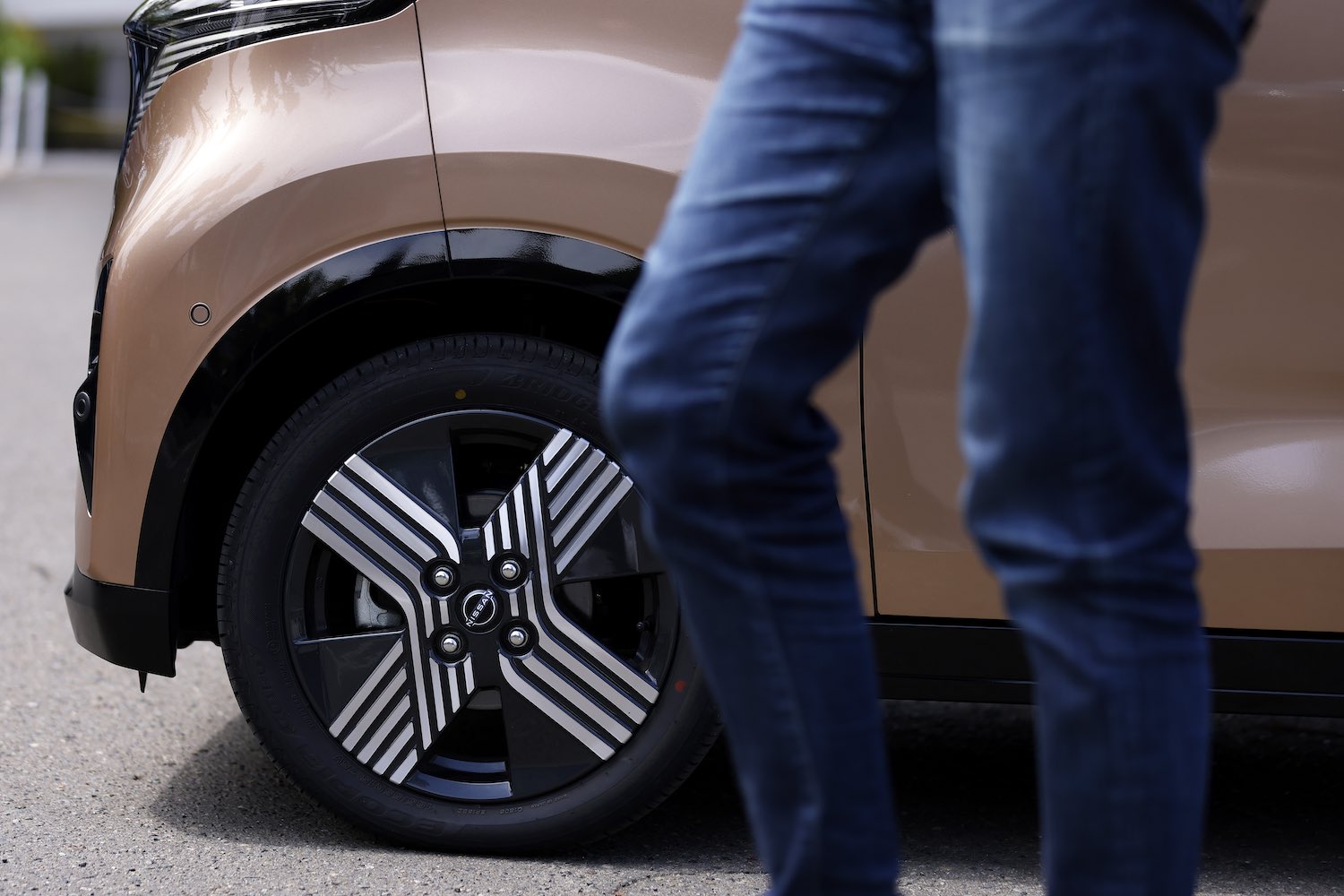 Closeup of the wheel and tire of a Nissan Sakura KEI-class EV, a person walking by in the foreground.