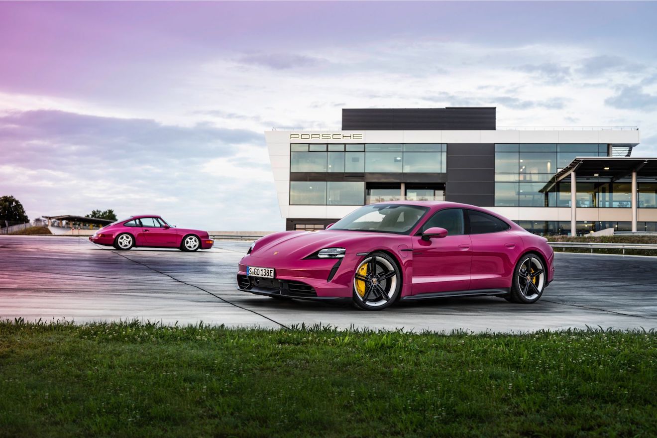 A Porsche Taycan in pink parked in front of a Porsche building. This is one of the few pink cars you can buy new.