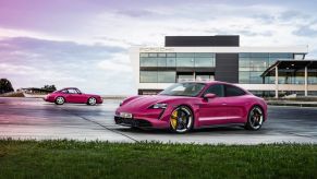 A Porsche Taycan in pink parked in front of a Porsche building. This is one of the few pink cars you can buy new.