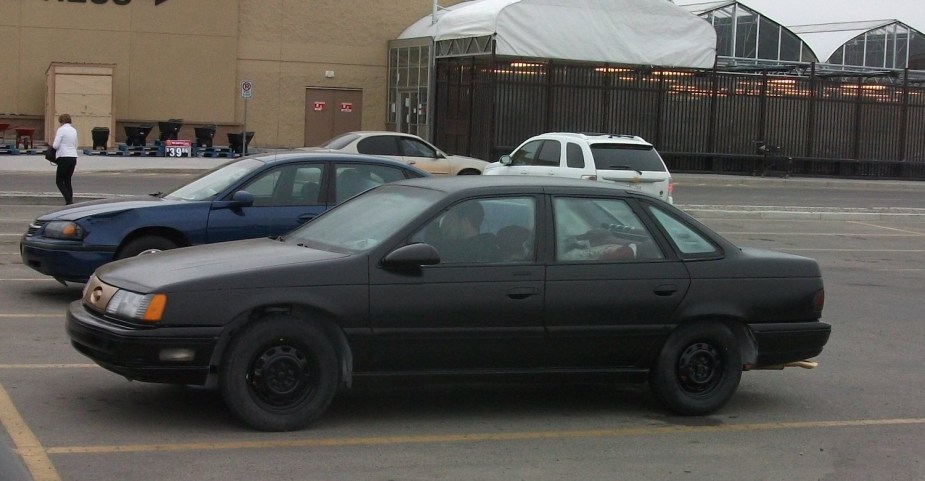 Blacked out 1986 Ford Taurus sedan in a parking lot, a building visible in the background.