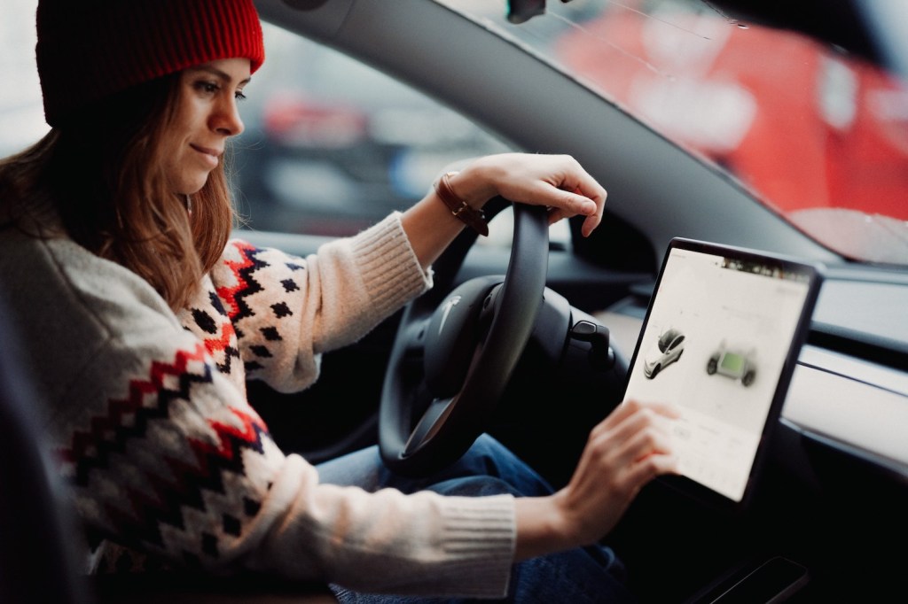 A woman uses a Tesla touchscreen to check advertised range, while sitting in an EV