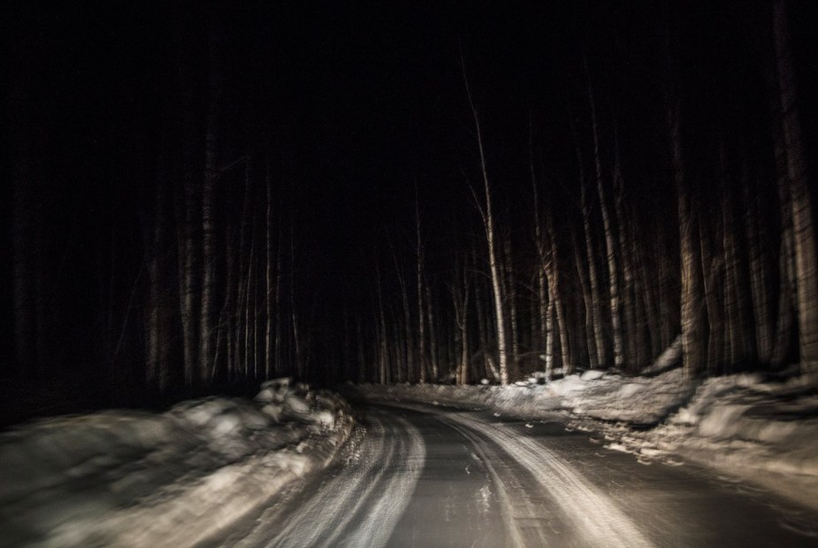 Headlights illuminate a snowy road at night, trees lining both sides.