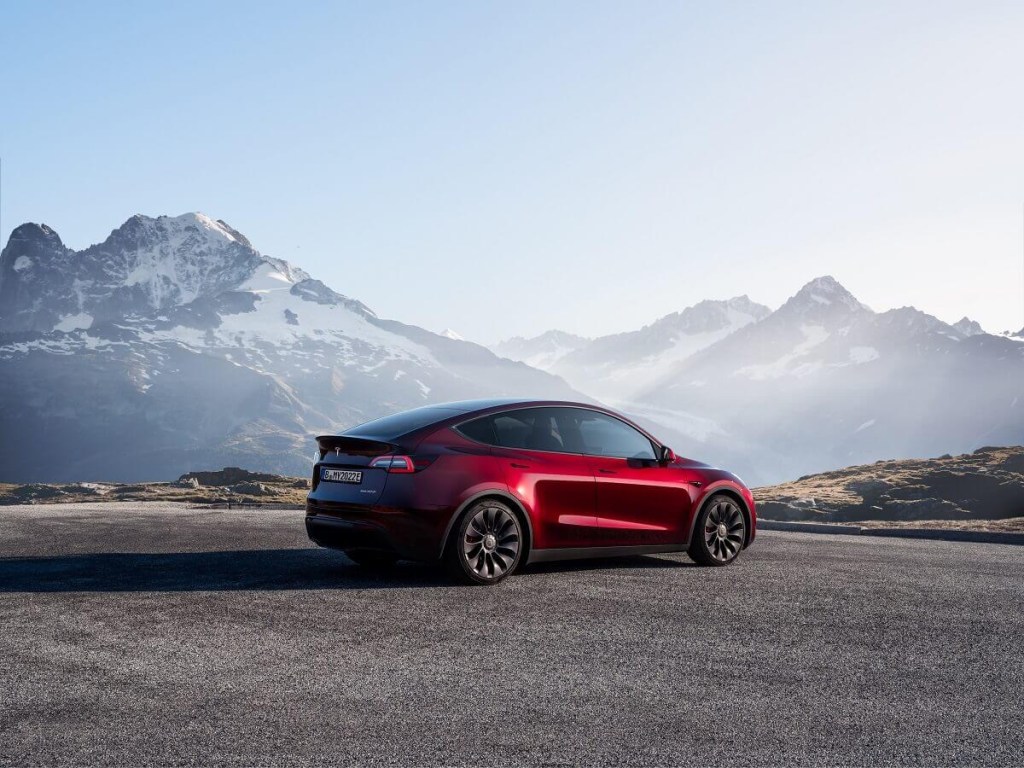 A red used 2023 Tesla Model Y Long Range, an EV SUV model with a Tesla price drop, parks near a mountain range.