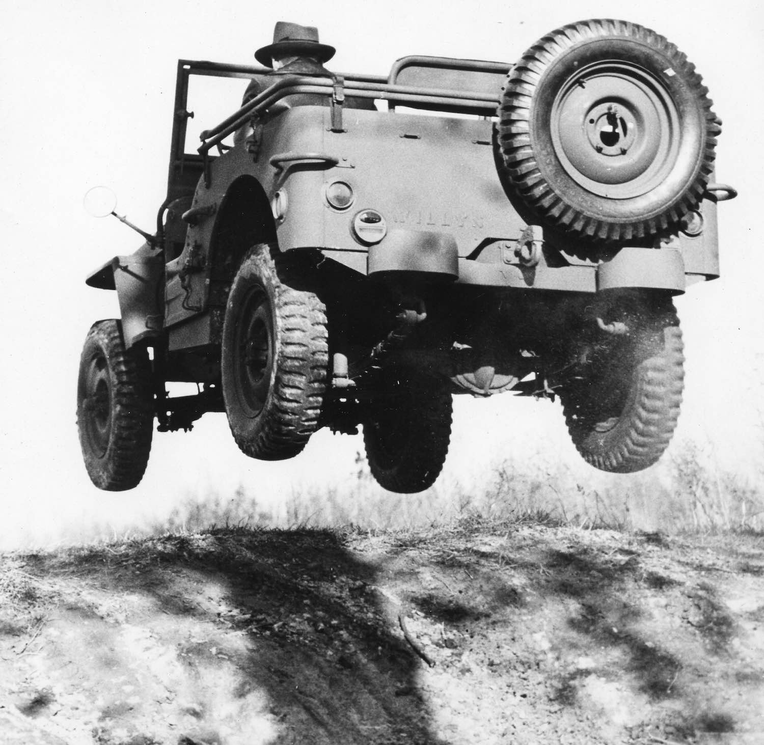 Black and white photo of a man in a fedora completing a jump across sand dunes in a Willys-Overland military GP Jeep prototype.