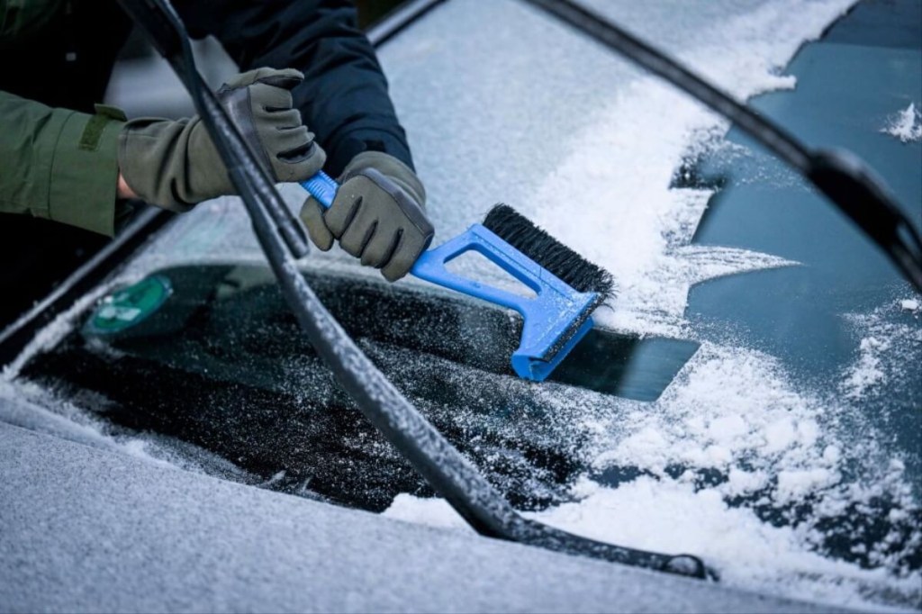 A car owner uses a car scraper and brush to clear snow from a car in the winterization process beyond winter tires and trickle charger use.