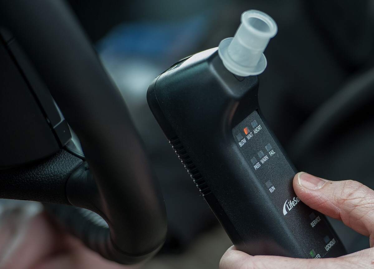 A hand holds a breath alcohol ignition interlock device (IID) next to a car steering wheel