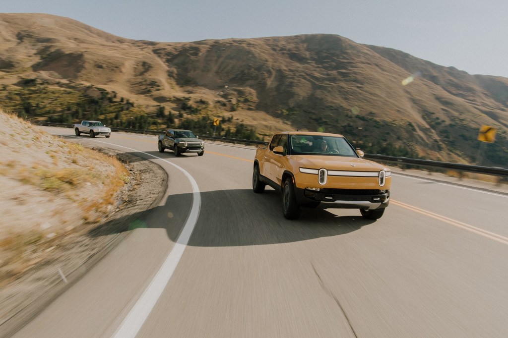 A line of three different colored Rivian R1Ts driving around a bend on a highway with mountains in the background